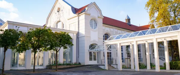 Elegantes, helles Gebäude mit klassischer Architektur, großen Fenstern und einem überdachten Eingangsbereich aus Glas und weißen Säulen, umgeben von Bäumen bei sonnigem Wetter.